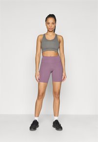 Γκρι αθλητικό crop top με ανοιχτοπράσινο λογότυπο, σετ με ψηλόμεσο λιλά σορτς και μαύρα αθλητικά παπούτσια. Ορατές λευκές κάλτσες αστραγάλου.