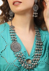 Collana dichiarativa argentata con più strati, caratterizzata da perline circolari e geometriche, abbinata a orecchini coordinati. Indossata sopra una maglietta teal.