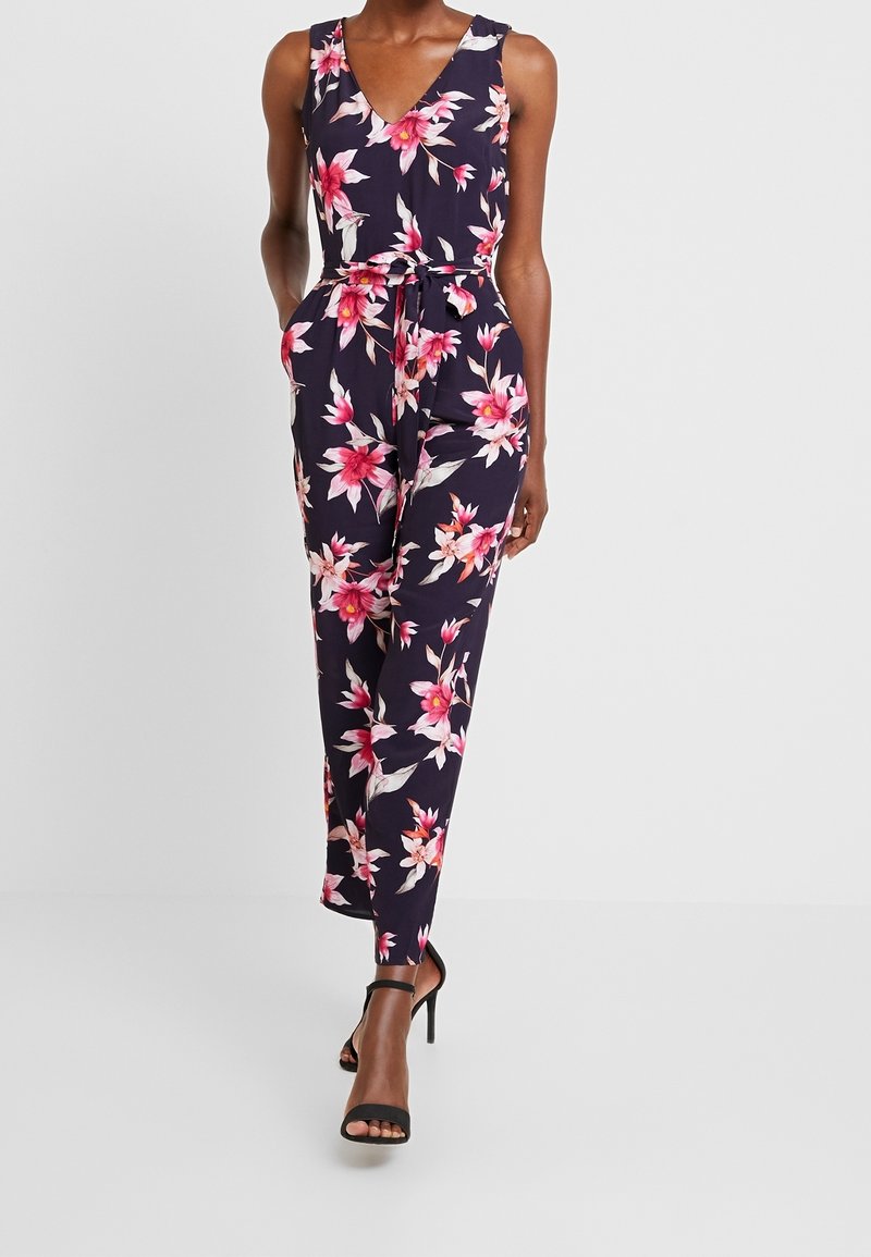 Combinaison fleurie bleu marine à fleurs roses, col en V, sans manches, taille ceinturée et jambes fuselées. Portée avec des sandales noires à talons.