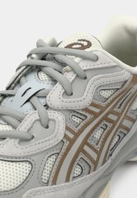 Γκρίζο αθλητικό παπούτσι με υφασμάτινη επάνω επιφάνεια με υφή, ανοιχτού γκρίζου κορδόνια και διακριτικές καφέ λεπτομέρειες. Ιδιαίτερο σχέδιο περιλαμβάνει στρωματοποιημένα, κυματιστά στοιχεία.