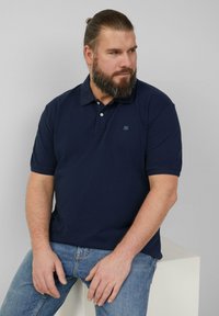 Granatowa koszulka polo z zapinanym kołnierzykiem, krótkimi rękawami i małym logo na lewej piersi. Noszona z jasnoniebieskimi dżinsami.