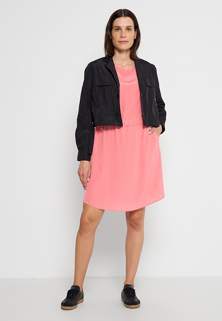 Schwarze kurze Jacke über einem pinken Kleid. Das Kleid ist glatt und hat eine lockere Passform, während die Jacke Vorder Taschen und Knöpfe hat. Schwarze Sneakers.
