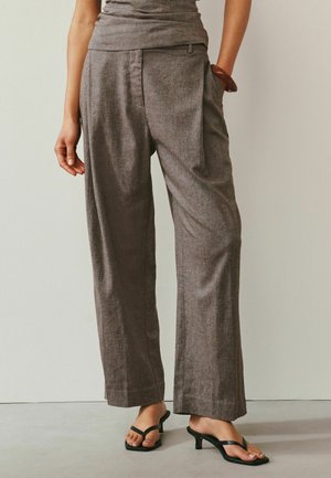 Woman wearing high-waisted, loose brown wide-leg trousers with pleats, paired with black heeled sandals, standing against a plain background.