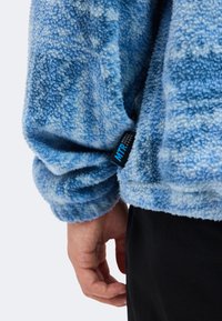 Άτομο φοράει μπλε fleece μανίκι με μοτίβο και μαύρη-μπλε ετικέτα "MTR" Mountain Training Range κοντά στον καρπό και μαύρο παντελόνι.