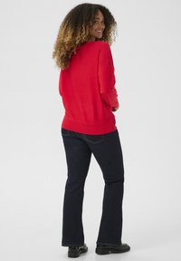 Punainen neule ribatulla helmallaan, yhdistetty tumma sinisiin leveälahkeisiin farkkuihin ja mustiin kenkiin, nähtynä takaa. Pehmeä rakenne, rento muotoilu.