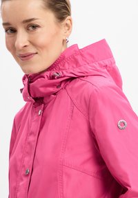 Rosa wasserdichte Jacke mit hohem Kragen, Druckknopfverschluss und silbernem Logo-Akzent am Ärmel. Glattes, strapazierfähiges Material.
