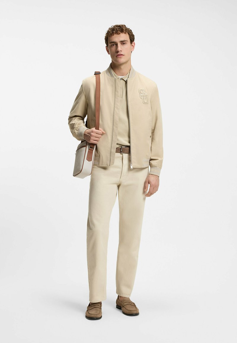 Homme portant une veste beige, un polo, un pantalon, une ceinture marron et des mocassins, avec un sac à bandoulière blanc et marron, debout sur un fond blanc.