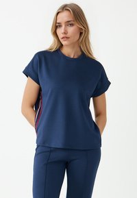 Kobieta z blond włosami, ubrana w granatową bluzkę z krótkim rękawem oraz spodnie, stoi z rękami w kieszeniach na tle białej ściany.