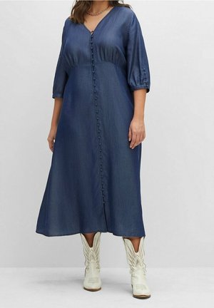 Vrouw draagt een lange blauwe jurk met knopen, driekwart mouwen en witte decoratieve cowboylaarzen, staand tegen een effen achtergrond.