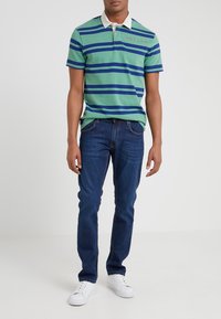 Grön- och blårandig polo skjorta med vit krage, med en röd logotyp på bröstet, tillsammans med mörkblå jeans och vita sneakers.