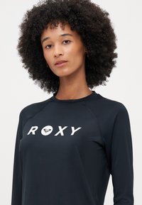 Melna garām piedurknēm izstrādājama ar lielu baltu "ROXY" logo un sirds grafiku. Izgatavota no gludas, vieglas materiāla ar raglan piedurknēm.
