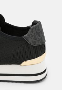 Czarny dzianinowy obcas sneakersa z teksturowaną szarą nakładką TPU i złotym akcentem nad białą podeszwą z czarnymi paskami na szarym tle.