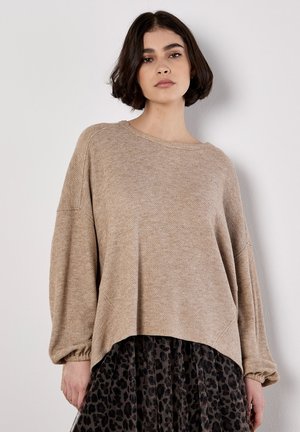 Pull en tricot beige à coupe ample, avec un col rond côtelé et des épaules tombantes. Associé à une jupe noire en tulle à imprimé léopard.