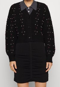 Cardigan noir en tricot avec un relief texturé et des embellissements en perles multicolores. En dessous, une jupe noire ajustée avec des détails froncés.
