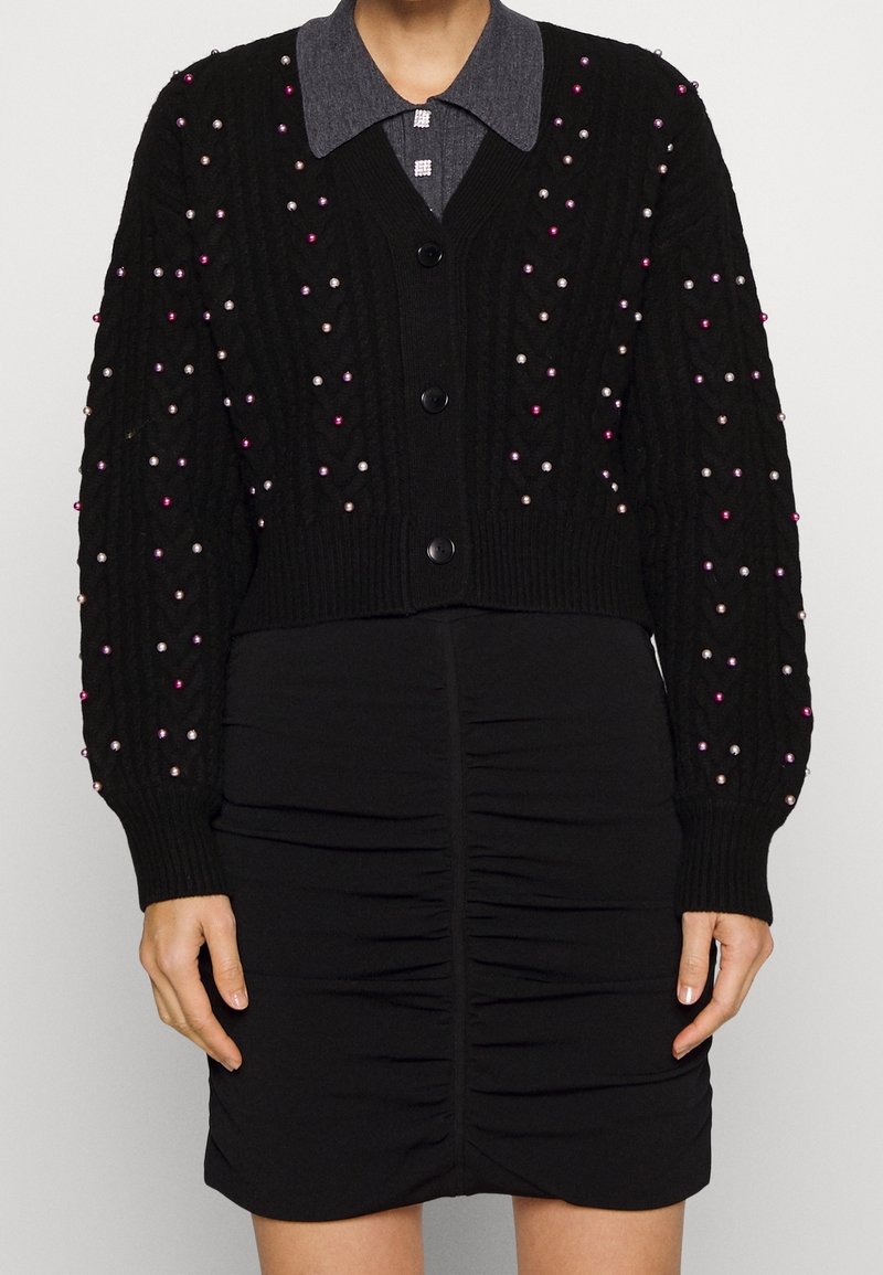Cardigan noir en tricot avec un relief texturé et des embellissements en perles multicolores. En dessous, une jupe noire ajustée avec des détails froncés.