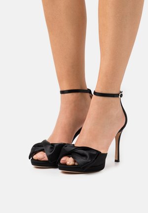 Sorte satin højhælede sko med en sløjfeaccent ved tåen, peep-toe design og en slank ankelrem med spænde.