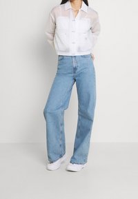 Giacca bianca trasparente corta con due tasche sul petto, abbinata a jeans a gamba larga color azzurro chiaro e sneakers bianche.