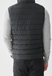 Black puffer vest with horizontal quilted pattern, high collar, and no sleeves, paired with a gray long-sleeve shirt and dark jeans.