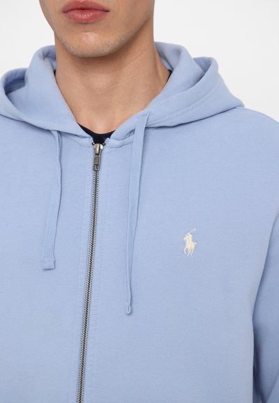 Sweatshirt azul claro com fecho éclair, feito de tecido macio. Apresenta um fecho na frente, cordões na capuz e um pequeno logotipo bordado no peito.