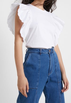 Femme portant un haut blanc sans manches à volants et un jean bleu taille haute, debout devant un fond uni.