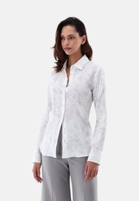 Camicia bianca a maniche lunghe con un sottile motivo floreale, dotata di colletto con bottoni, realizzata in tessuto leggero e con bottoni blu a contrasto.