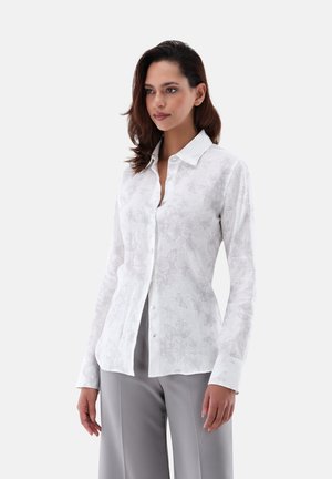 Camicia bianca a maniche lunghe con un sottile motivo floreale, dotata di colletto con bottoni, realizzata in tessuto leggero e con bottoni blu a contrasto.