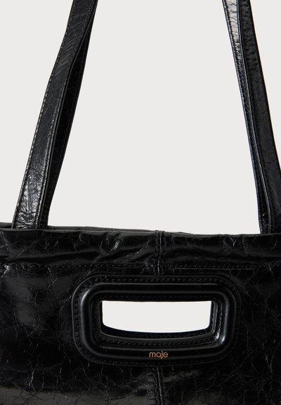 Sac à main en cuir texturé noir avec deux bretelles d’épaule et une poignée découpée rectangulaire étiquetée "maje" en petites lettres dorées.