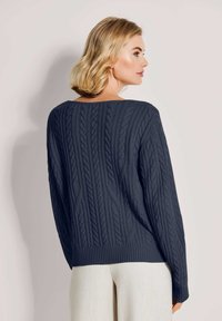 Blonde Frau, die einen navyblauen Rundhalspullover aus Zopfmuster und hellbeige Hosen trägt, seitlich vor einem schlichten hellen Hintergrund steht.