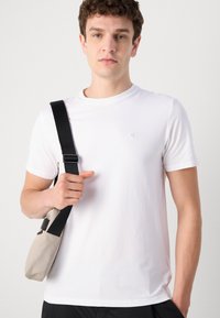 Witte katoenen T-shirt met een ronde hals en korte mouwen, met een klein grijs logo op de borst. Zwarte band houdt een beige tas vast.