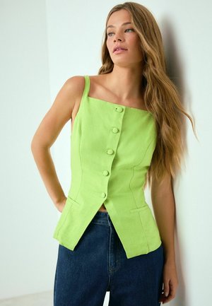 Sieviete, kas atbalstās pret sienu, tērpusies dzīvžodziņzaļā bezroku topā ar pogām un tumši zilās džinsa biksēs, skatās uz sāniem.