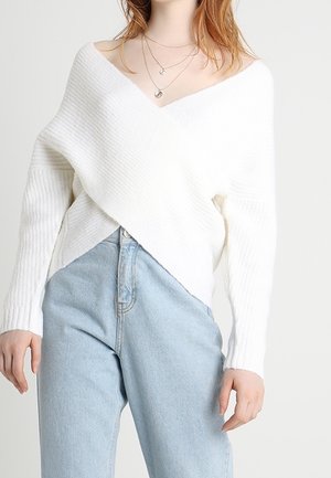 Frau trägt einen weißen, gerippten V-Ausschnitt-Strickpullover, in hoch taillierte hellblaue Jeans gesteckt, accessoriert mit geschichteten silbernen Halsketten.