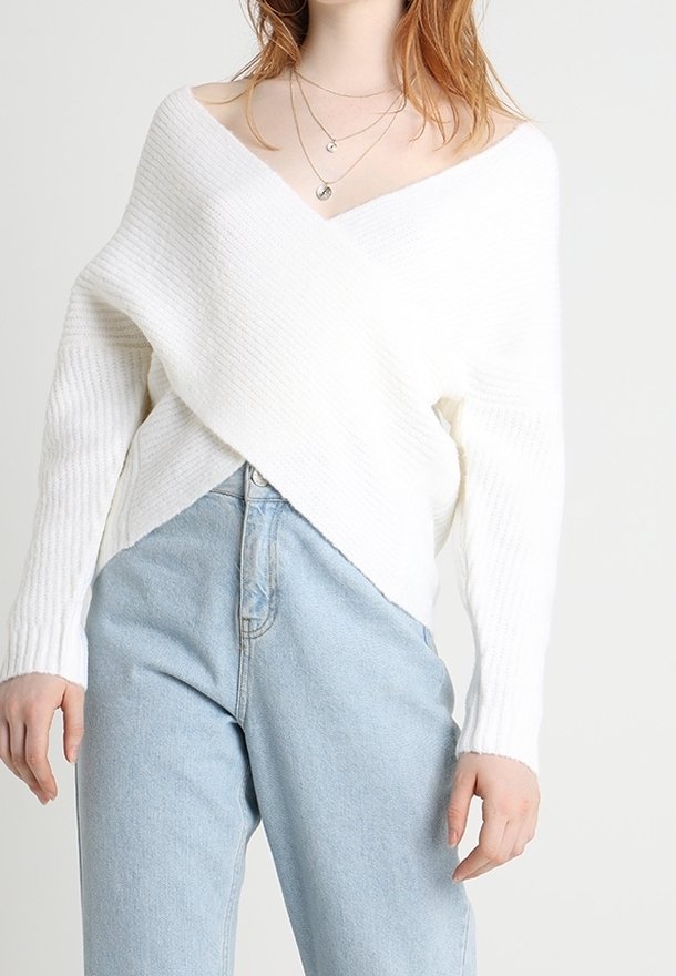 Femme portant un pull en maille blanc côtelé à col en V, rentré dans un jean taille haute bleu clair, accessoirisée de colliers argentés superposés.