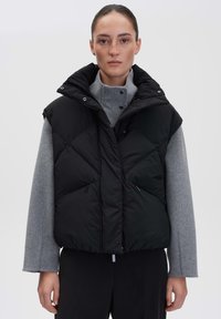 Schwarze Pufferweste mit hohem Kragen, Rautenmuster und Reißverschlusstaschen, getragen über einem grauen Pullover. Matte Oberfläche und übergroßes Design.