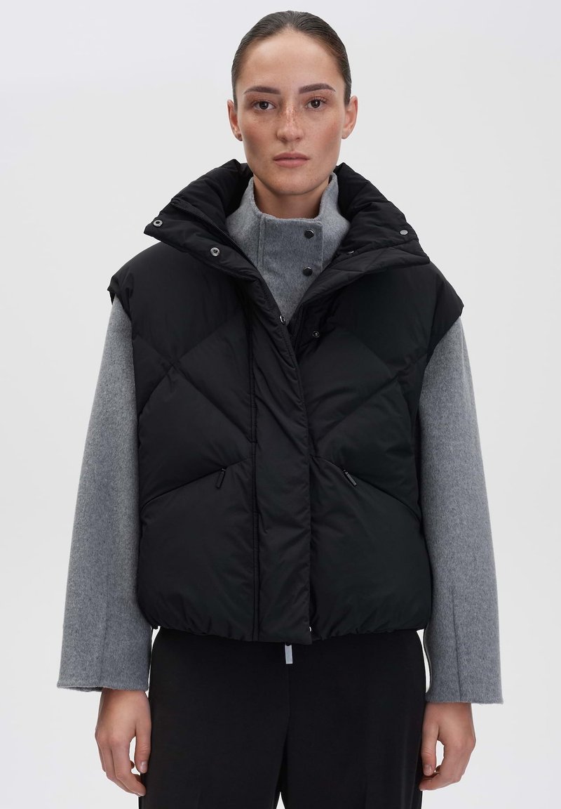 Schwarze Pufferweste mit hohem Kragen, Rautenmuster und Reißverschlusstaschen, getragen über einem grauen Pullover. Matte Oberfläche und übergroßes Design.