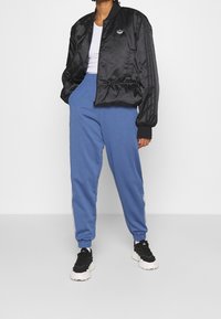 Svart satin bomberjacka med ribbade ärmar, tillsammans med blå sweatpants med vita sidostreck. Mörka skor fulländar outfiten.