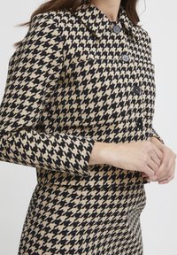 Houndstooth-mustriga jakk musta ja beeži värviskeemiga. Omab kraed, nuppe ja kitsast lõiget. Tekstuurne kangas on nähtav.