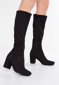 Botas negras hasta la rodilla, hechas de un tejido suave con textura brillante, tacones cuadrados y cierre de cremallera lateral. Diseño liso y ajustado.