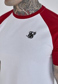 Valge T-särk punaste raglan-särkide varrukatega, millel on rindkeres must tikandiga logo. Valmistatud siledast puuvillaste materjalidest.