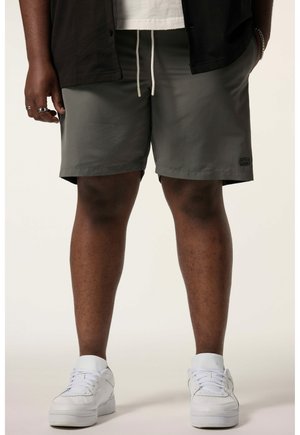 Mężczyzna w szarych spodenkach z sznurkiem, białych trampkach, czarnej koszulce, stojący z rękami w kieszeniach na jasnym tle.