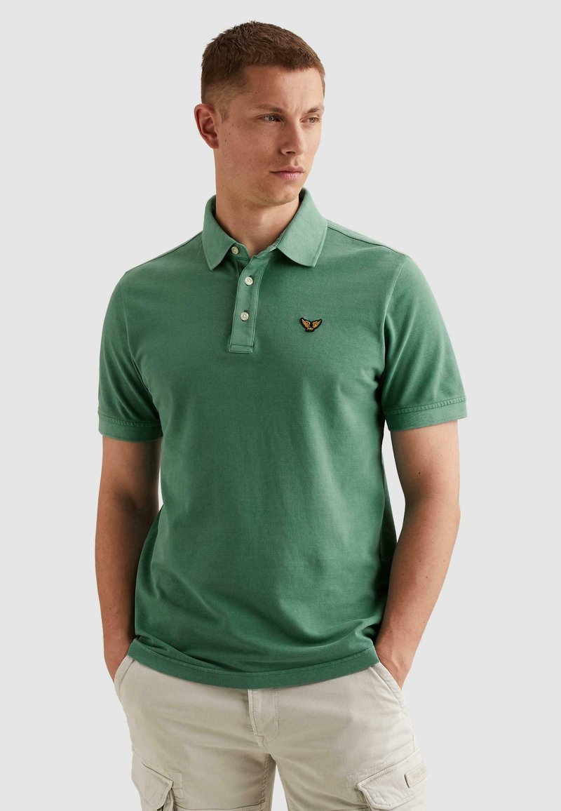 Jeune homme aux cheveux courts portant un polo vert à manches courtes avec un petit emblème brodé, un pantalon cargo beige, les mains dans les poches.
