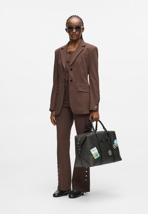 Ruskea räätälöity puku, johon kuuluu istuva bleiseri ja leveälahkeiset housut. Musta kuviollinen laukku, jossa on matkatarroja. Mustat aurinkolasit ja kengät luovat kontrastia.