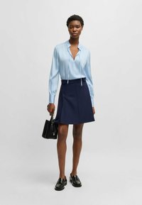 Blusa azzurra chiara a maniche lunghe con scollatura a V e dettaglio con fiocco abbinata a una gonna pantalone blu navy. Il modello tiene una piccola borsa nera ed indossa mocassini neri.
