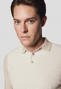 Beżowa koszulka polo z teksturowanej tkaniny, z wyraźnym kołnierzykiem, trzema guzikami z przodu oraz subtelnym wzorem w kratkę.