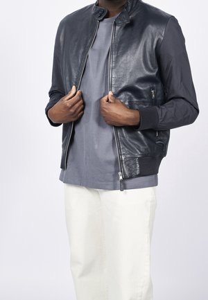 Homme portant une veste bomber en cuir bleu marine sur un t-shirt gris et un pantalon blanc, tenant les bords de la veste avec les deux mains au niveau de la poitrine.