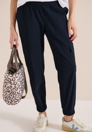 Kvinde iført sorte cargobukser og hvide sneakers, der holder en beige og brun leopardmønstret håndtaske ved sin side.