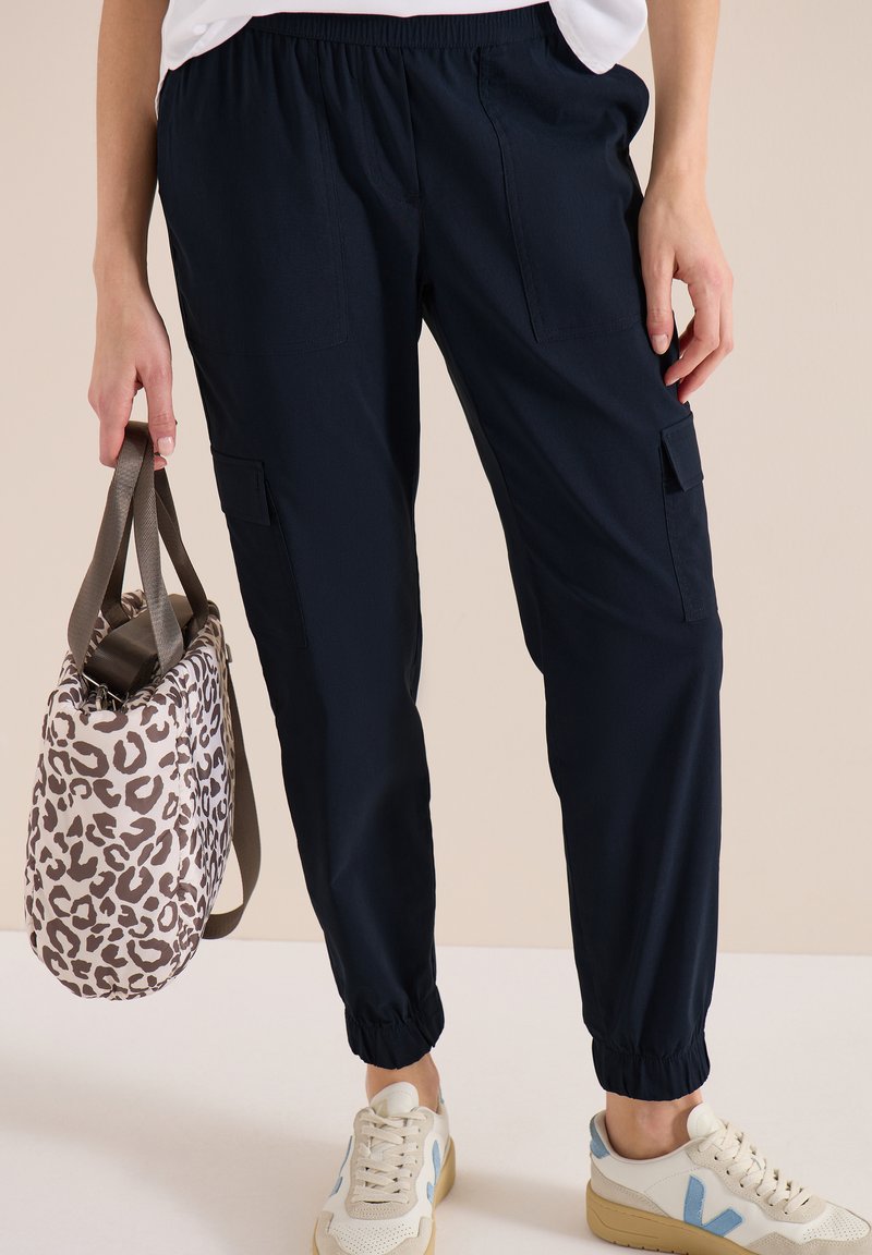 Femme portant un pantalon cargo noir et des baskets blanches, tenant à ses côtés un sac à main beige avec imprimé léopard marron.