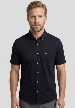 Man met licht haar en baard, die een zwarte korte mouwen overhemd met kraag en een klein logo op de borstzak draagt.