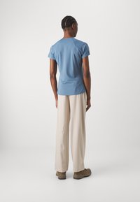 Camicia blu a maniche corte con scollo rotondo, design aderente e texture liscia, abbinata a pantaloni ampia di colore chiaro e scarpe marroni.