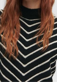 Pull en maille noir avec des rayures blanches en chevron, doté d'un col montant et d'une texture douce, encadrant de longs cheveux ondulés.