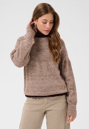 Ung kvinde med langt brunt hår iført en strikket beige sweater, beige bukser og farverige hængende øreringe mod en ensfarvet baggrund.